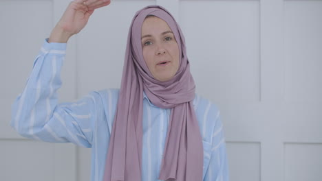 Looking-at-the-camera-muslim-girl-calling-friends-from-apartments-using-a-video-link-waving-with-hand-sitting-on-a-sofa-in-a-stylish-room-in-front-of-computer-screen-having-video-conversation